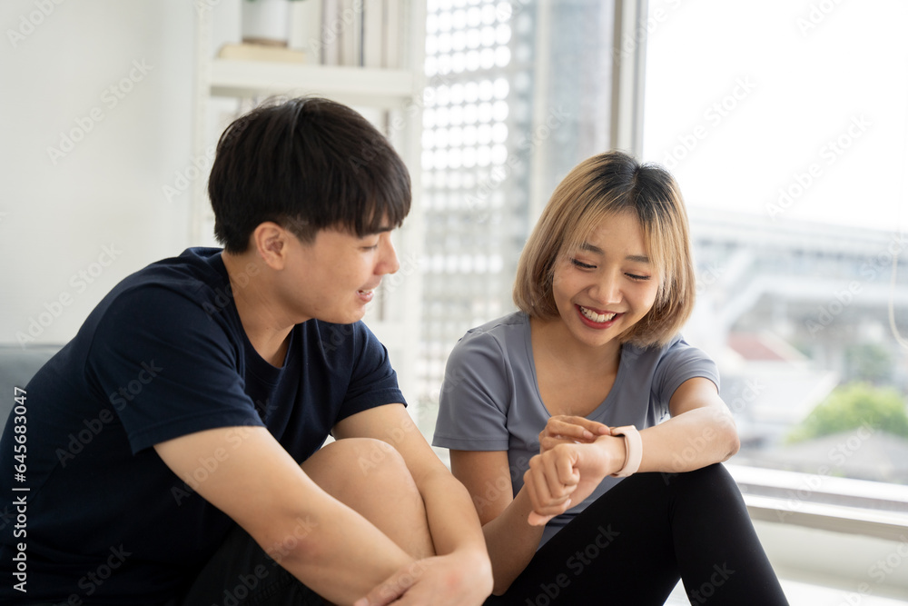 Happy asian people sitting on living room together. Woman Showing detail in smart watch with smiling while rest from exercise at home. Happy asian people sitting on living room together. Woman Showing detail in smart watch with smiling while rest from exercise at home.