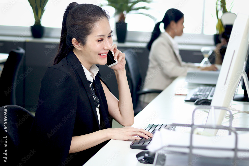 Happy smiling Asian woman officer sitting, talking on mobile phone at office desk with blurred busy colleague background, modern fashionable female businesswoman beautiful working at workplace. Happy smiling Asian woman officer sitting, talking on mobile phone at office desk with blurred busy colleague background, modern fashionable female businesswoman beautiful working at workplace.