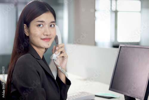 Beautiful Asian woman operator officer talking with customer by using landline phone at working desk in office with busy working colleagues blurred background. Smiling female receptionist at workplace