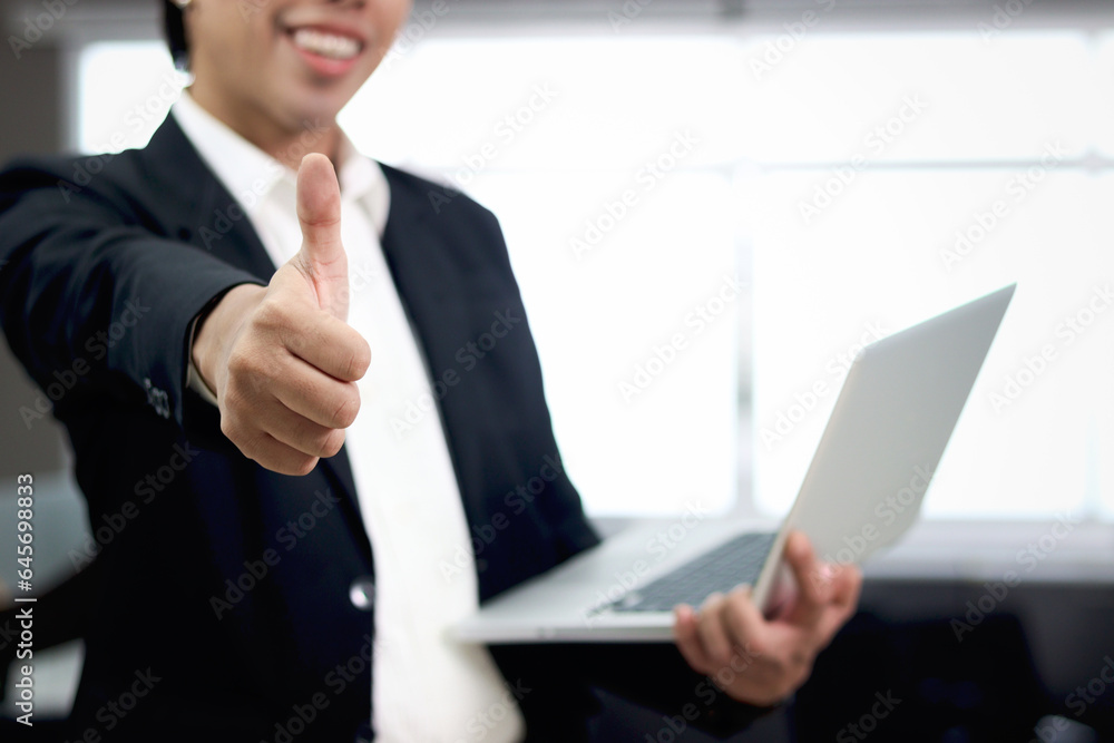 Happy smiling Asian businessman in black suit giving thumb up to camera while holding laptop computer, standing inside office building, confident male officer working at workplace. Happy smiling Asian businessman in black suit giving thumb up to camera while holding laptop computer, standing inside office building, confident male officer working at workplace.