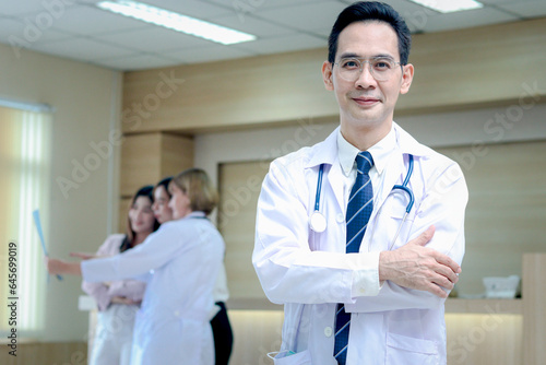 Portrait of smiling senior Asian male doctor in workwear with stethoscope  standing with arms crossed in clinic hospital  looks at camera and medical team working with patient as blurred background.