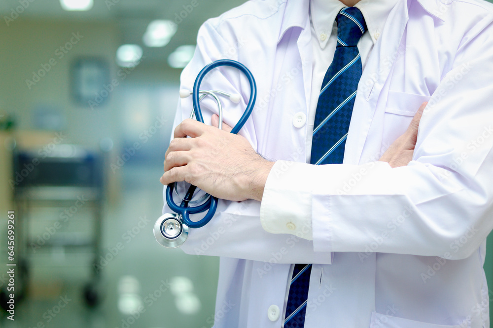 Confident male doctor in white workwear holding stethoscope standing with arms crossed in corridor of medical clinic, doctor working at hospital. medical and health care concept. Confident male doctor in white workwear holding stethoscope standing with arms crossed in corridor of medical clinic, doctor working at hospital. medical and health care concept.
