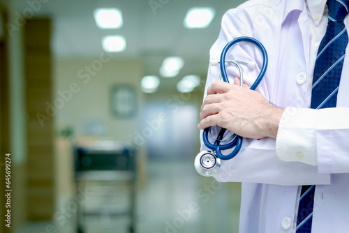 Fotografie Confident male doctor in white workwear holding stethoscope standing with arms crossed in corridor of medical clinic, doctor working at hospital