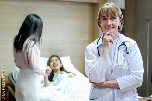 Portrait of senior female doctor in workwear with stethoscope standing and thinking and taking decisions with hand on chin in recovery room at clinic hospital with patient lying on bed as background.