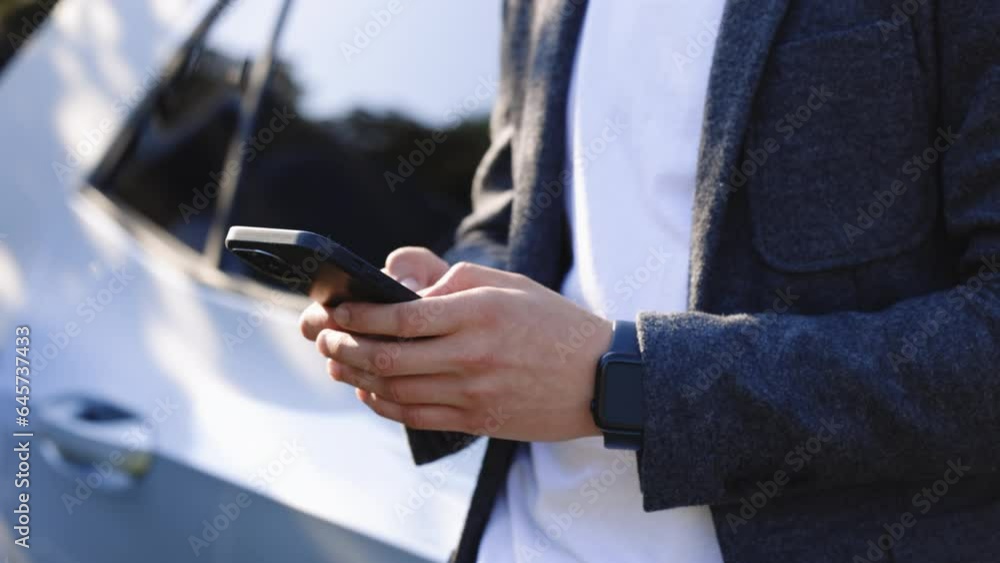 Man holding smartphone while charging car at electric vehicle charging station, closeup. Electric car charging, businessman standing in background and waiting Man holding smartphone while charging car at electric vehicle charging station, closeup. Electric car charging, businessman standing in background and waiting