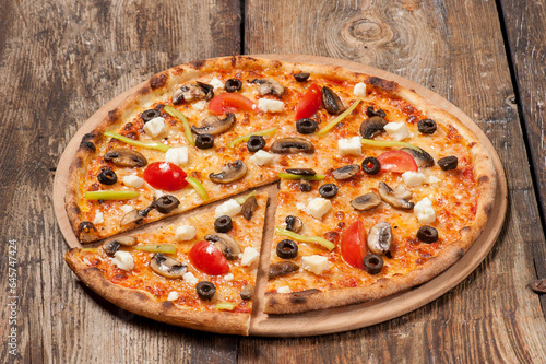 Studio shot of a hand made pizza with a cut slice with mushrooms, tomato, green peppers, olives and feta cheese on beautiful weathered wooden table