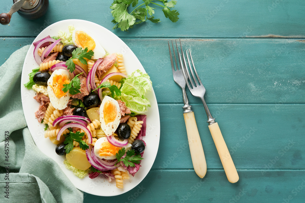 Tuna salad with pasta, eggs, potatoes, olives, red onions and sauce in white plate on old turquoise wooden rustic table background. Nicoise salad. French cuisine. Top view. Flat lay. Tuna salad with pasta, eggs, potatoes, olives, red onions and sauce in white plate on old turquoise wooden rustic table background. Nicoise salad. French cuisine. Top view. Flat lay.