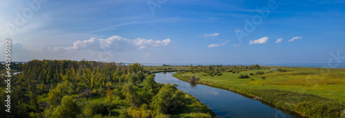 Early autumn panoramic aerial view of a winding river cutting through forests and grassy areas under a blue sky that has clouds that are white and fluffy and also diffuse.
