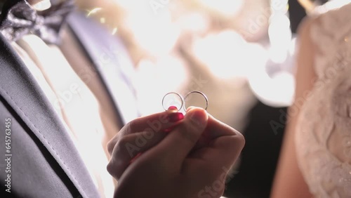 A close-up of newlyweds holding two wedding rings together in soft, shimmering light. The elegant scene features gentle flickering, lens flares, and warm backlighting, creating a romantic and slightly