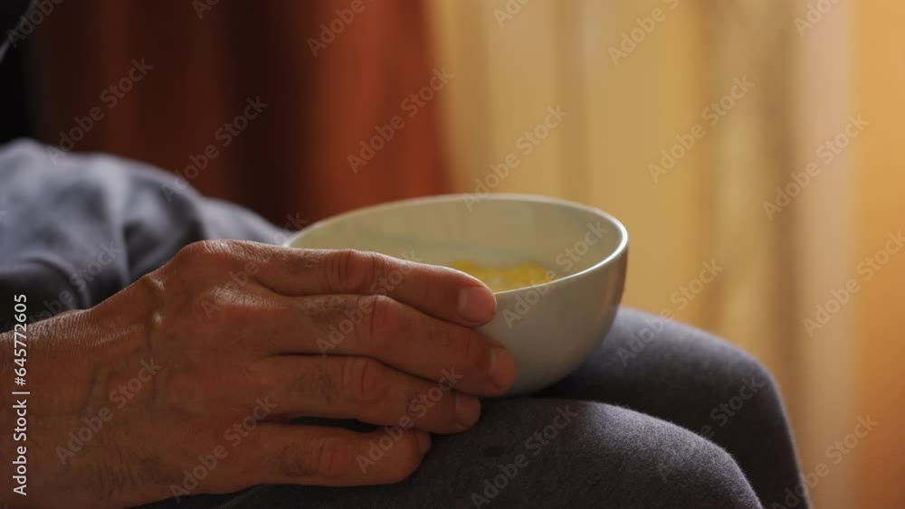 The older man's hands hold the bowl in his lap during breakfast. The older man's hands hold the bowl in his lap during breakfast.