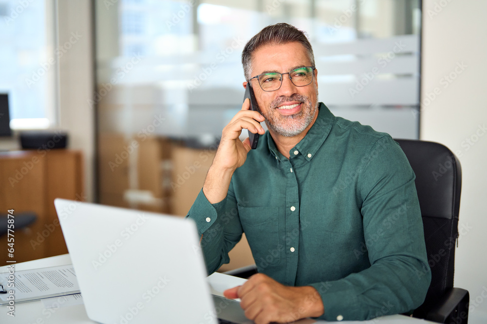 Smiling mid aged businessman making call on cellphone using laptop at work. Happy busy mature old professional business man manager talking on phone working communicating in office looking aside. Smiling mid aged businessman making call on cellphone using laptop at work. Happy busy mature old professional business man manager talking on phone working communicating in office looking aside.
