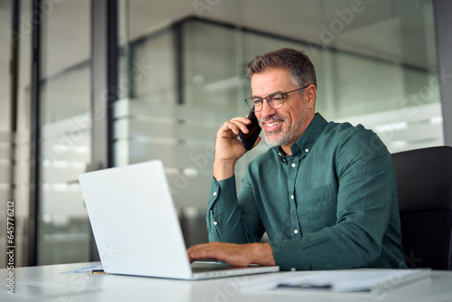 Busy smiling older middle aged business man professional expert or entrepreneur making phone call speaking with client communicating on cellphone using laptop computer sitting at desk in office.