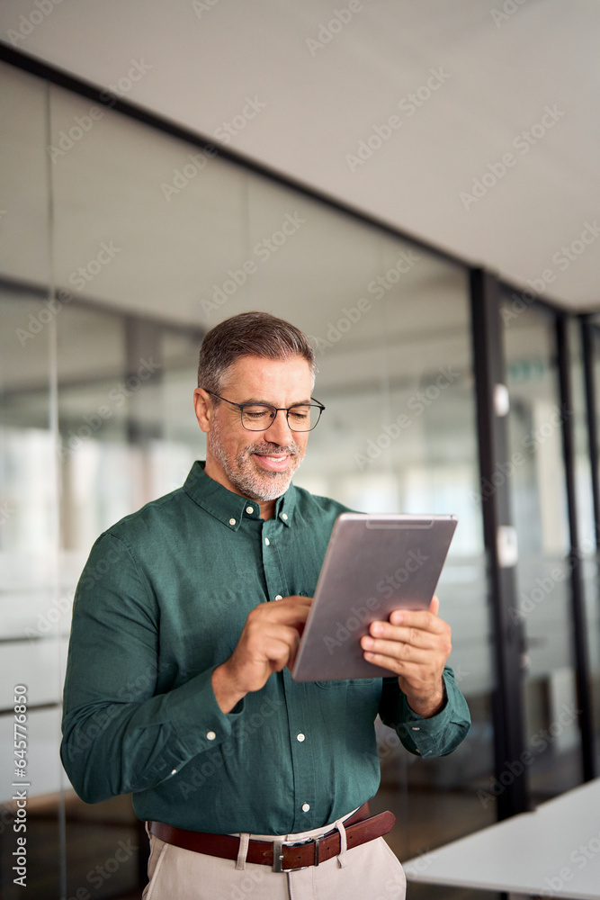 Smiling busy middle aged business man investor using tablet computer, mature older businessman executive manager looking at tab satisfied with finance trading market standing in office. Vertical Smiling busy middle aged business man investor using tablet computer, mature older businessman executive manager looking at tab satisfied with finance trading market standing in office. Vertical