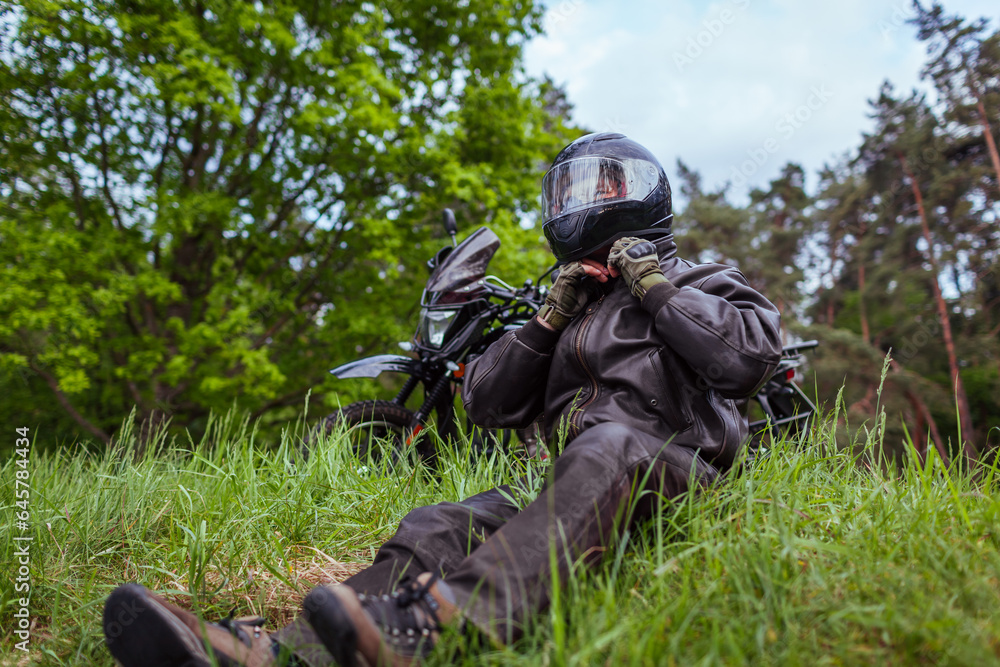 Biker unfastening helmet after riding motobike in summer forest. Man wearing leather clothes outdoors. Biker unfastening helmet after riding motobike in summer forest. Man wearing leather clothes outdoors.