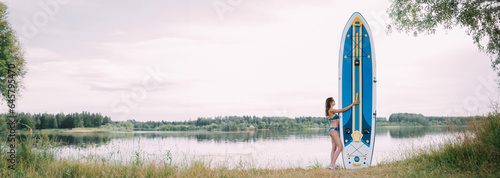 A young woman with an open swimsuit stands with a sapboard on the shore of the lake. Stand Up Paddle. SUP touring photo