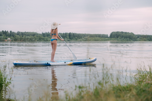 A young woman with an open swimsuit swims on a SUP board on a picturesque lake. Substitute the paddle. A beautiful, slender girl is engaged in sap surfing on the calm water of a picturesque pond.