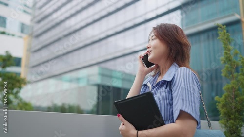 Young asian woman talking on smartphone with friend rejoicing smiling holding tablet at outdoors. Young businesswoman or university studen talking on smartphone at city view photo