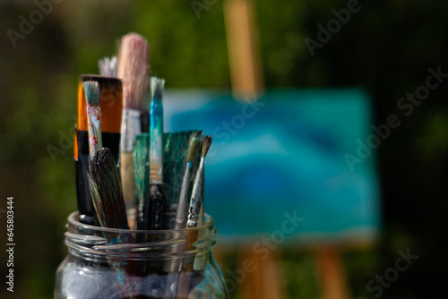 Many brushes in a glass jar with an easel with a painted canvas behind
