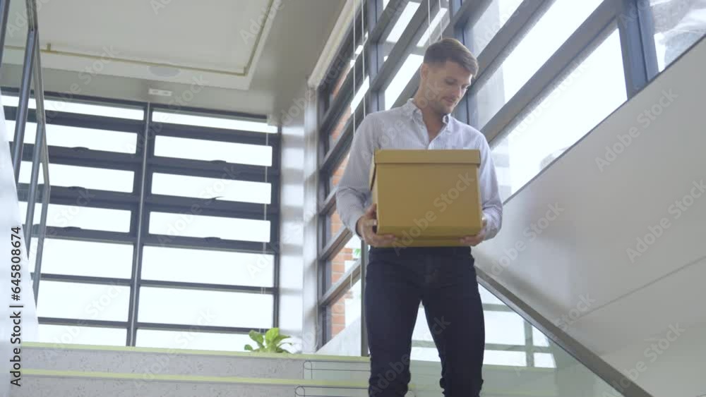 Caucasian man hold box and walk down from stair to lower level or floor look like he quit or lose job and take his things to go out from office. Caucasian man hold box and walk down from stair to lower level or floor look like he quit or lose job and take his things to go out from office.