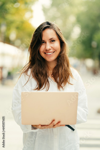 Cheerful young smiling woman is walking down a street while holding her laptop in summer.