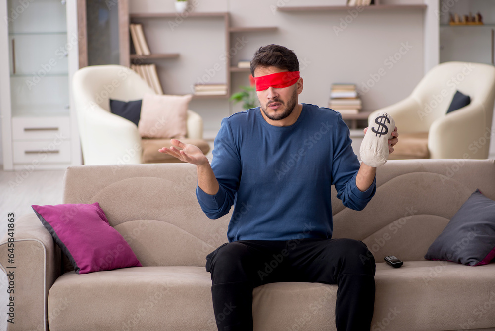 Young blindfolded man holding moneybag at home Young blindfolded man holding moneybag at home