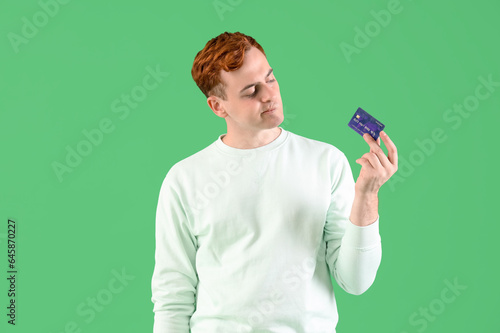 Thoughtful young redhead man with credit card on green background