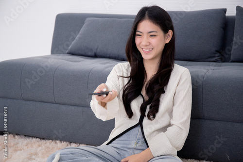 Beautiful young asian woman sitting using remote control with television for watching series with cozy and happiness in living room at home, girl looking cinema or movie on tv, lifestyles concept.