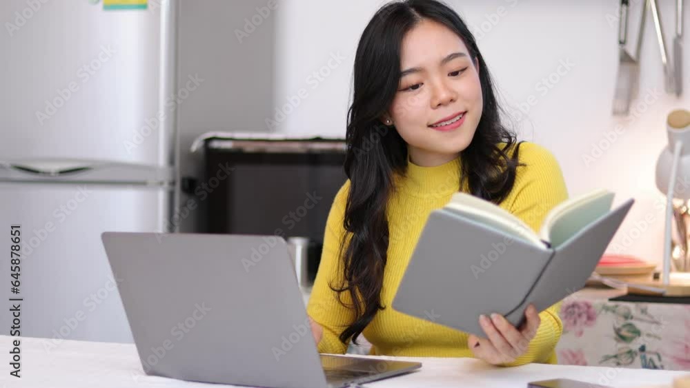 Young Asian woman working on laptop in kitchen at home, she is taking cooking class online and writing recipe notes from internet. Young Asian woman working on laptop in kitchen at home, she is taking cooking class online and writing recipe notes from internet.