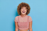 Smiling curly-haired girl in pink spotted top against blue background laughing out with closed eyes, good mood concept, copy space
