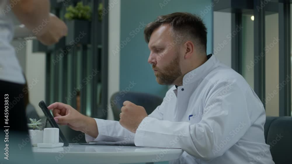Male doctor sits in modern clinic cafe. Healthcare specialist works on digital tablet, surfs the Internet and have meal. Canteen workers brings order to medic. Medical staff in modern hospital. Male doctor sits in modern clinic cafe. Healthcare specialist works on digital tablet, surfs the Internet and have meal. Canteen workers brings order to medic. Medical staff in modern hospital.