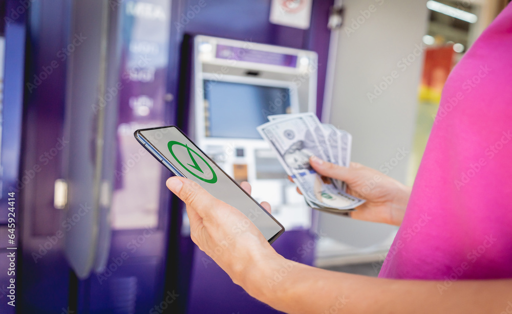 Young woman using the smart phone for cardless withdrawing the cash near the ATM Young woman using the smart phone for cardless withdrawing the cash near the ATM