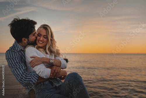 Handsome man holding beautiful woman in his arms and kissing her while enjoying the sunset. Couple in love sitting at the beach during their honeymoon.