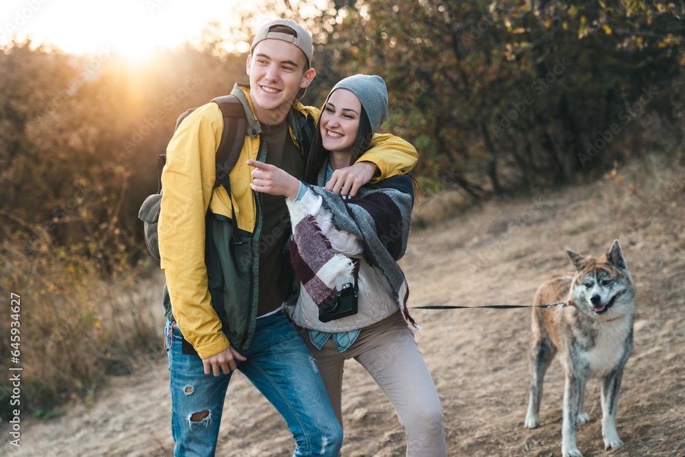 Young guy and girl smiling and walking a dog in nature. He is hugging her and holding a dog leash. Sunny day in Autumn.