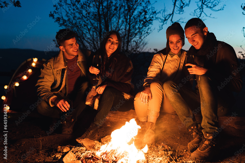 In forest, night time, four best friends are sitting by fire and having a good talk. They are looking at campfire and smiling. In forest, night time, four best friends are sitting by fire and having a good talk. They are looking at campfire and smiling.