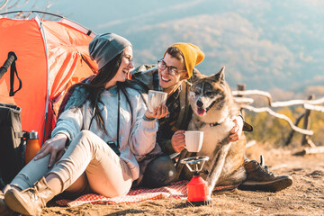 Photo of a happy young couple and their dog camping in the mountains on a beautiful autumn day and drinking a cup of tea outdoors.