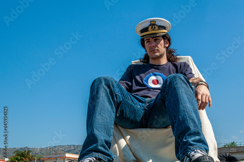 Giovane uomo vestito casual visto in prospettiva dal basso seduto con cappello della marina mercantile italiana e sfondo del cielo azzurro
