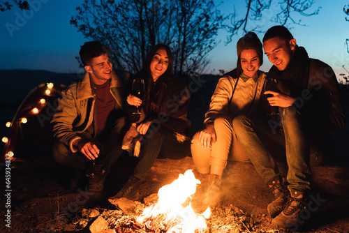 In forest, night time, four best friends are sitting by fire and having a good talk. They are looking at campfire and smiling.