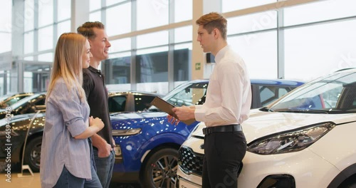 Good looking salesman showing some cars to couple in car dealership Slow motion Guy consulting family to buy vehicle Business Conversation Luxurious autos. Car dealer showing car to couple in showroom