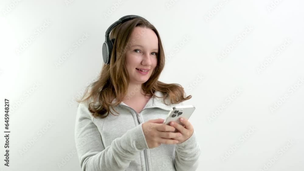 girl with her finger taps on headphones large professional headphones listen to music check quality in hands holds white smartphone mobile phone internet wi-fi on white background laughs. smile girl with her finger taps on headphones large professional headphones listen to music check quality in hands holds white smartphone mobile phone internet wi-fi on white background laughs. smile