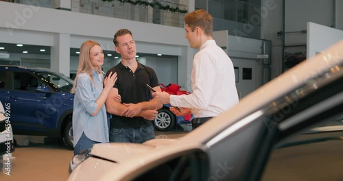 handsome male shop assistant in formal clothes helping caucasian young family in choosing buying purchasing new car at automobile dealer shop store. skilled confident male manager talking to clients