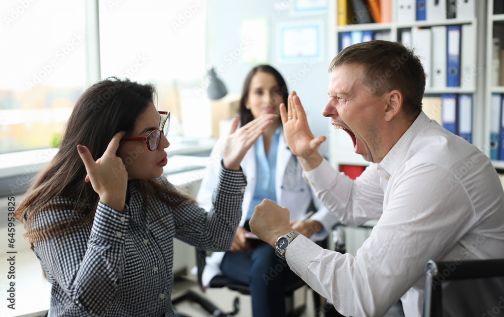 Focus on angry married couple doc appointment in clinic office. Shocked woman showing to screaming man fool gesture. Loud and emotional quarrel in clinic. Family planning center concept Focus on angry married couple doc appointment in clinic office. Shocked woman showing to screaming man fool gesture. Loud and emotional quarrel in clinic. Family planning center concept