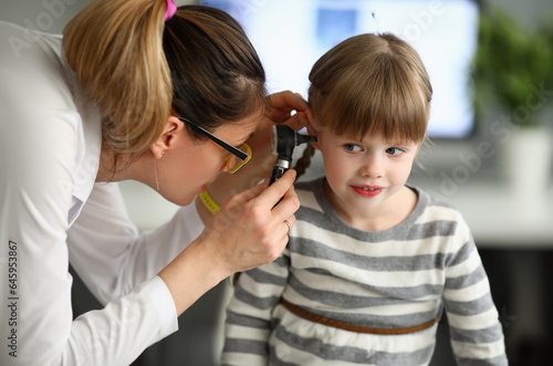 Portrait of joyful girl sitting in clinic office and looking away with happiness. Professional therapist watching in child ear with special utility. Health checkup concept