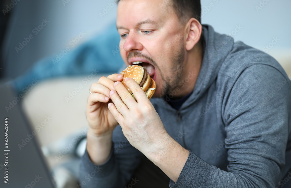 Portrait of male sitting indoors and eating big tasty hamburger full of mischievous calories and harmful cholesterol. Person consuming portion of junk food Portrait of male sitting indoors and eating big tasty hamburger full of mischievous calories and harmful cholesterol. Person consuming portion of junk food