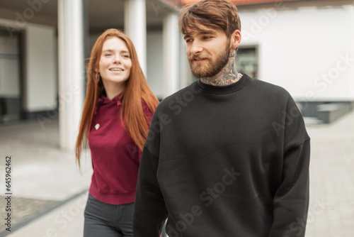 Happy beautiful young fashionable couple redhead beautiful woman with smile and brutal hipster handsome guy with tattoo in trendy fashion pullover walking in the city