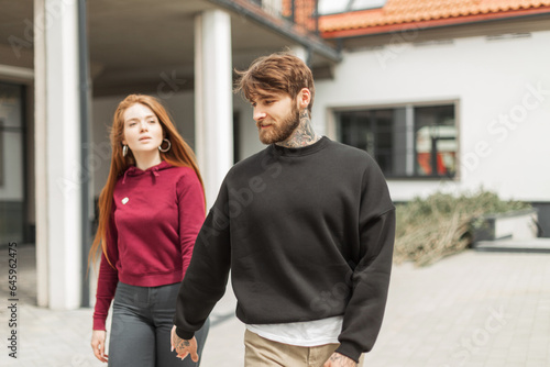 Beautiful fashionable couple in a stylish hoodie walks in the city. Handsome hipster man with a beard and a beautiful red-haired girl are walking together and holding hands