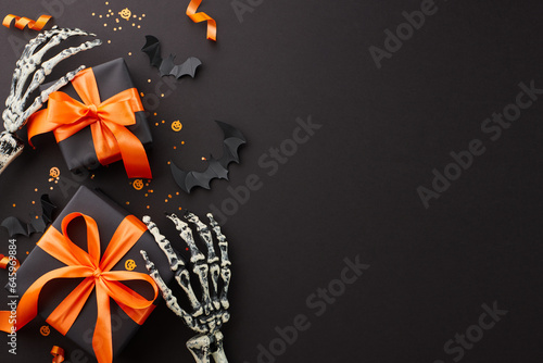 It's time for Halloween savings. Top view composition of skeleton hands, satin bows, black gift boxes, spooky bats, confetti on black background with advert space