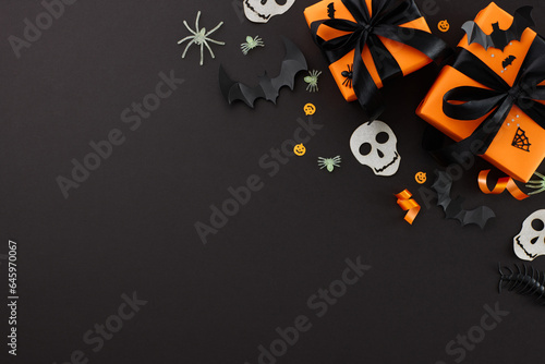 Spooktacular Halloween savings. Top view flat lay of gift boxes, satin bows, paper skulls, scary insects, shiny confetti on black background with commercial placeholder