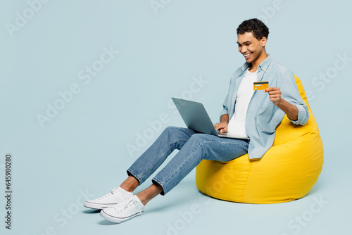 Full body young man of African American ethnicity wear shirt casual clothes sit in bag chair using mobile cell phone hold credit bank card shopping online book tour isolated on plain blue background.