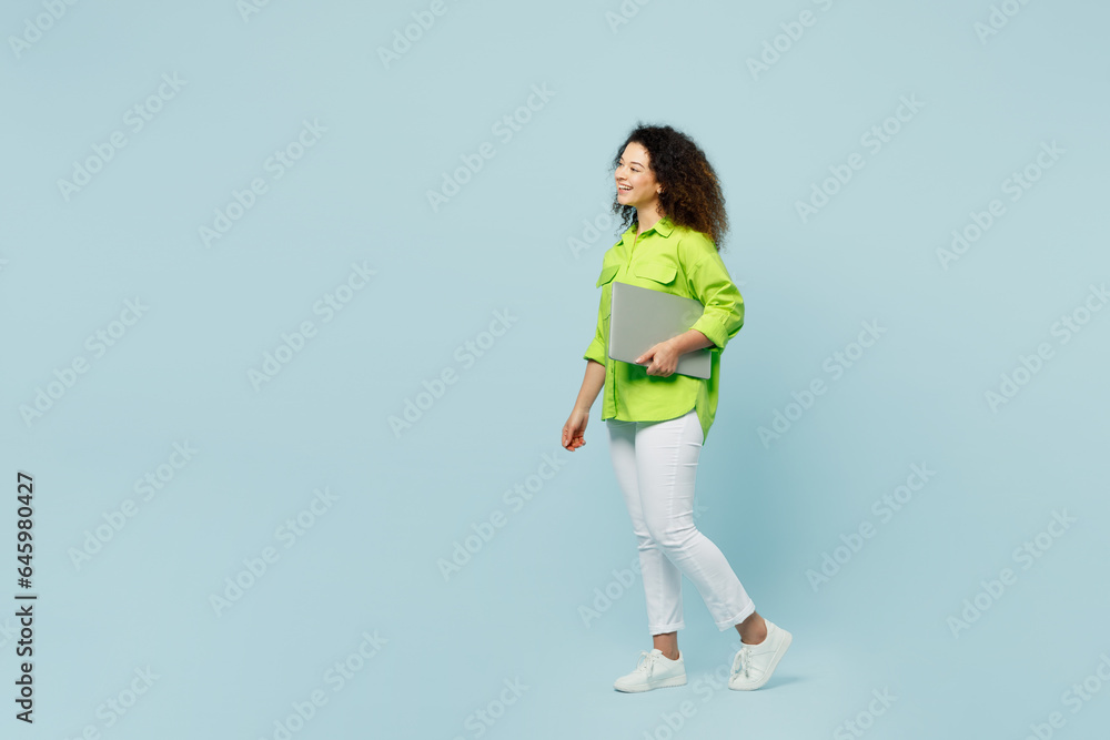 Full body side profile view young IT latin woman wear green shirt casual clothes hold closed laptop pc computer isolated on plain pastel light blue cyan background studio portrait. Lifestyle concept. Full body side profile view young IT latin woman wear green shirt casual clothes hold closed laptop pc computer isolated on plain pastel light blue cyan background studio portrait. Lifestyle concept.