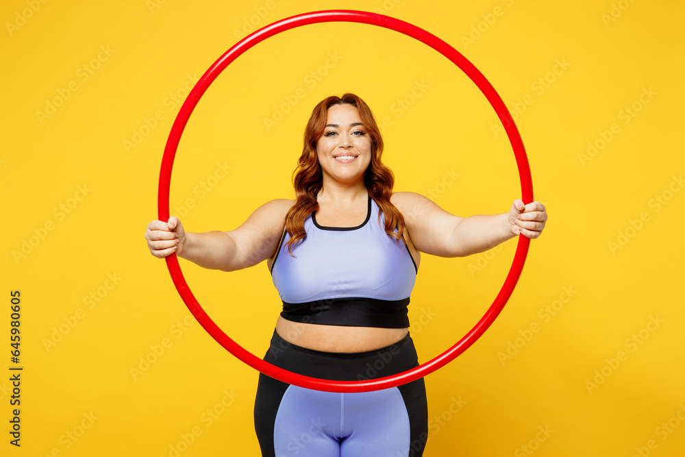 Young smiling happy fun chubby overweight plus size big fat fit woman wear blue top warm up training look through hula hoop isolated on plain yellow background studio home gym. Workout sport concept. Young smiling happy fun chubby overweight plus size big fat fit woman wear blue top warm up training look through hula hoop isolated on plain yellow background studio home gym. Workout sport concept.
